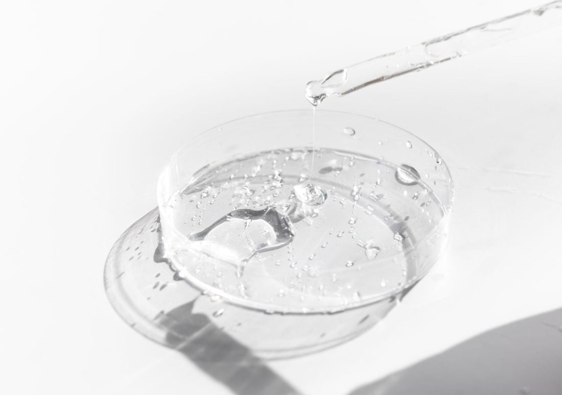 Translucent, slightly viscous essence droplets on a clear glass dish beside a small serum vial